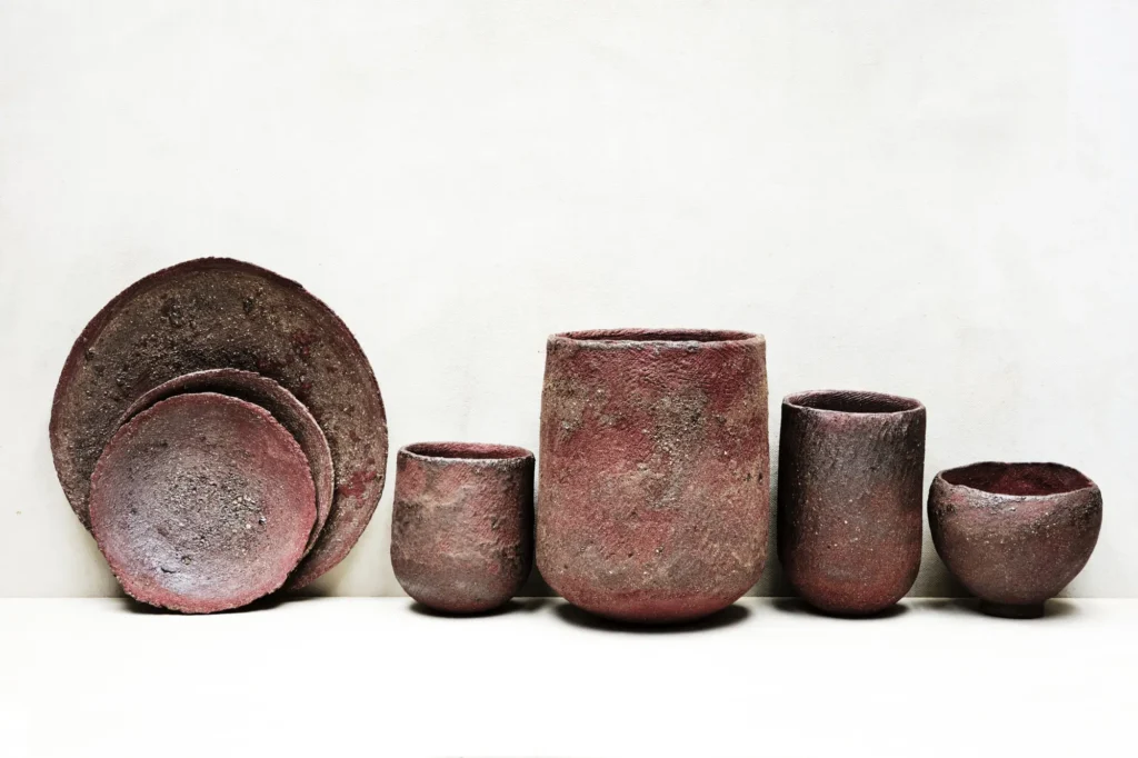 Dunkelrötliches Stoneware-Geschirr mit rauer Struktur und deutlicher Patinierung, aufgereiht vor hellem Hintergrund