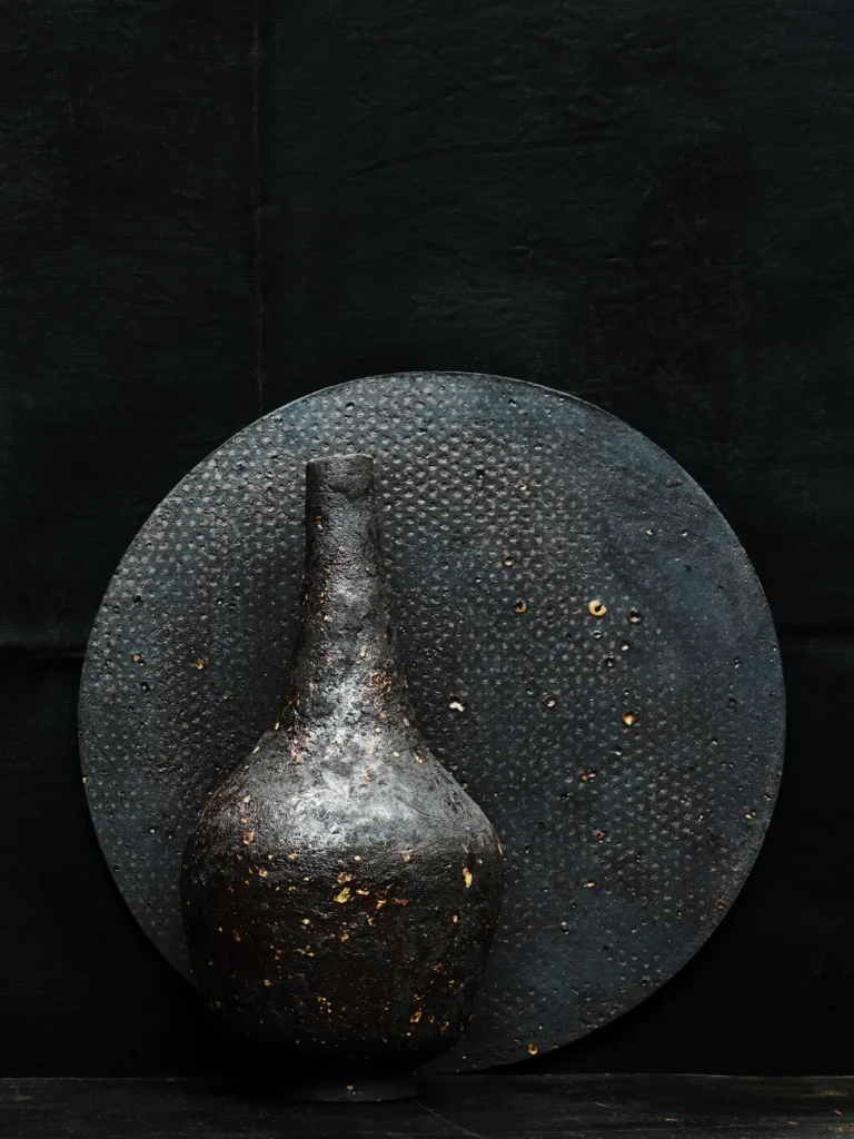 Schwarzer Stoneware-Teller und -Vase mit löchriger Streetdirt-Struktur, aufgenommen vor schwarzem Hintergrund