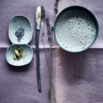 Stillleben mit adaptogenem Moon-Milk-Getränk, Lavendelzweigen und Gewürzen in handgeformten Schalen auf violettem Tuch, arrangiert von Dietlind Wolf