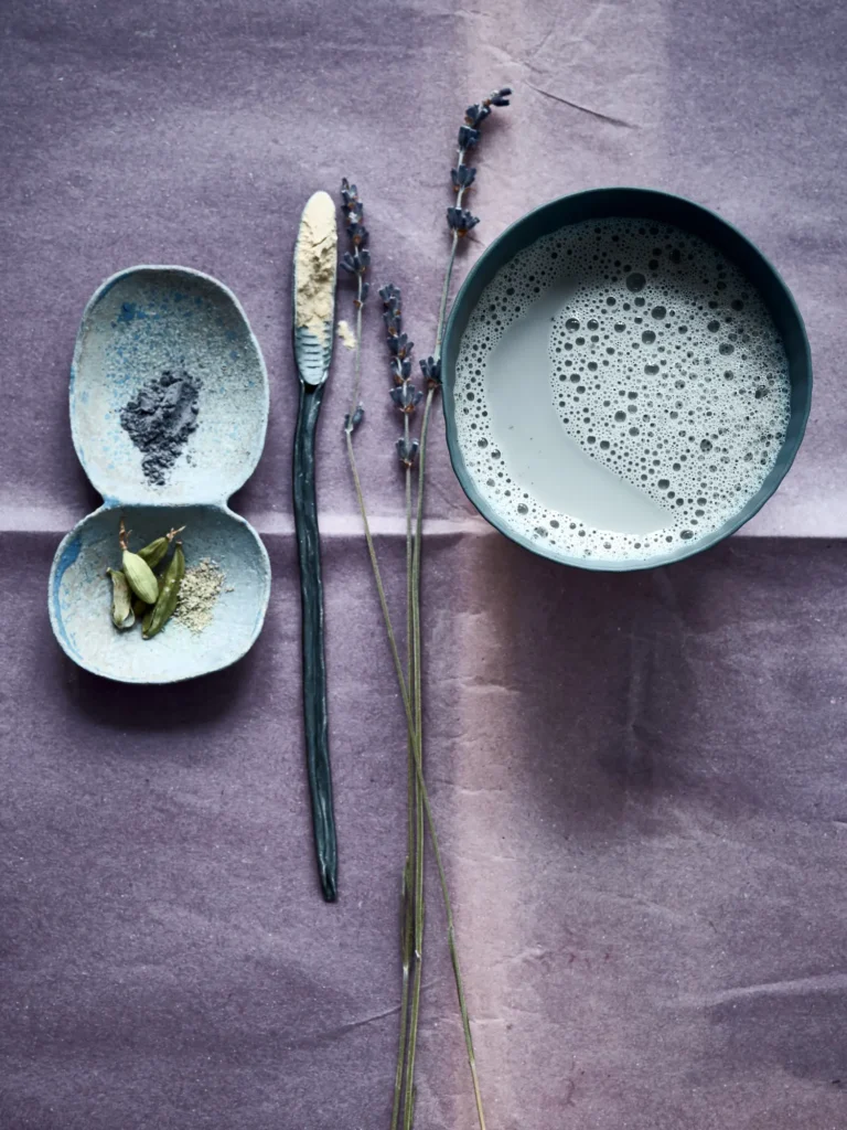 Stillleben mit adaptogenem Moon-Milk-Getränk, Lavendelzweigen und Gewürzen in handgeformten Schalen auf violettem Tuch, arrangiert von Dietlind Wolf