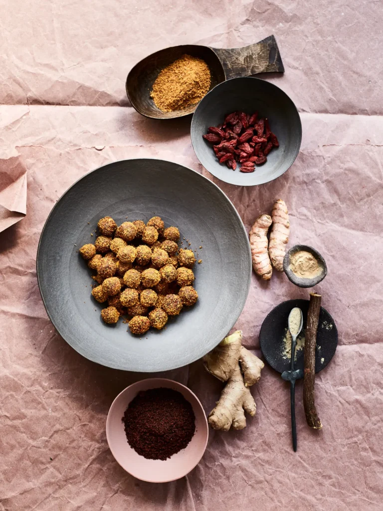 Graue Schale mit runden Kugeln, braune Gewürzpulver in kleinen Schalen, getrocknete Beeren, gelbe Wurzelstücke, frischer Ingwer auf rosa Papier mit Holzlöffeln"