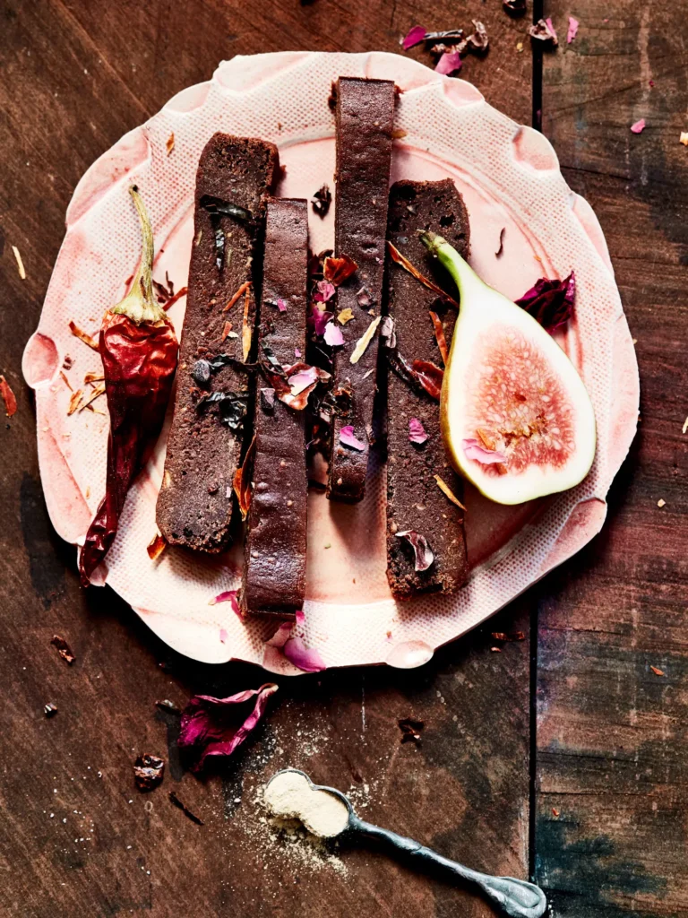 Dunkle Kuchenstreifen auf rosa Keramikteller mit Rosenblättern, getrockneter Chilischote und einer aufgeschnittenen Feige von Dietlind Wolf, auf dunkler Holzoberfläche mit Pulverlöffel