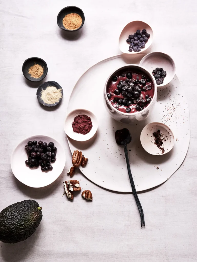 Dunkle Beeren-Bowl in heller Keramikschale, umringt von kleinen Schälchen mit verschiedenen Pulvern, getrockneten Beeren, Pekannüssen und einer Avocado von Dietlind Wolf, auf hellem Leinenhintergrund