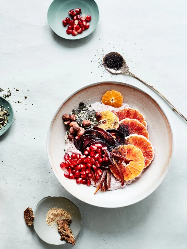 Helle Keramikschale mit Porridge, getoppt von Blutorangenscheiben, Granatapfelkernen, Nüssen und dunklen Fruchtstreifen, arrangiert von Dietlind Wolf, umringt von kleinen Schälchen mit Granatapfelkernen, Kernen und Pulver sowie einem gefüllten Löffel auf hellem Grund