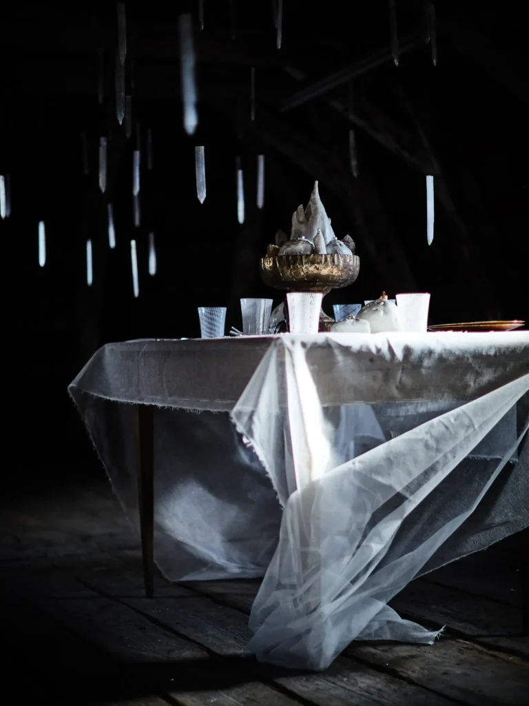 Dunkel inszenierter Tisch mit transparentem Tuch, Gläsern und Schale; darüber schwebend herabhängende, längliche Kristalle, dramatisches Licht und starke Schatten.“
