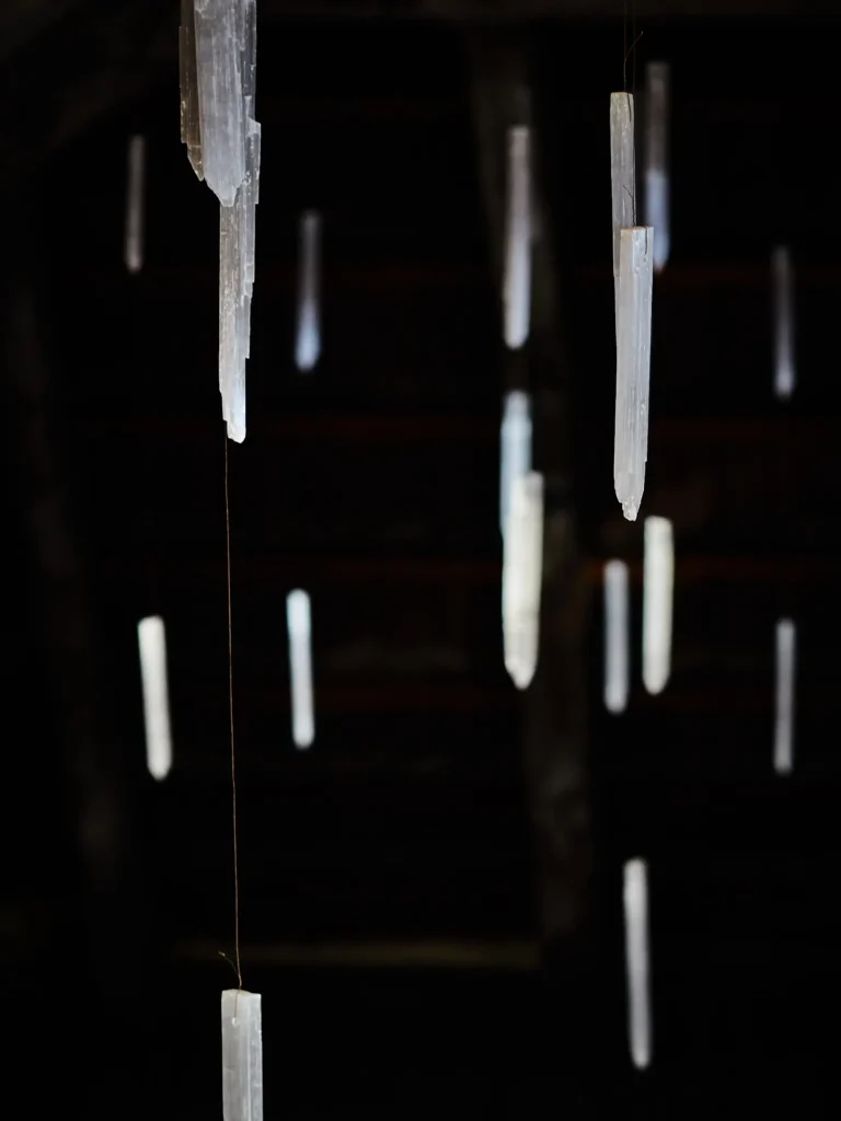 Mehrere längliche, transparente Kristalle hängen an feinen Fäden frei im dunklen Raum; gestaffelte Tiefenschärfe, ruhige Komposition, kühles Licht vor schwarzem Hintergrund.