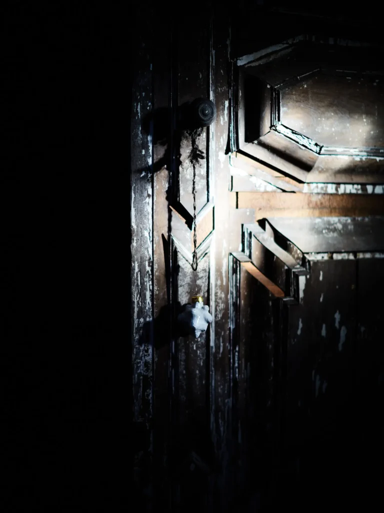 Dunkle, verwitterte Holztür mit abblätternder Farbe und kleinem hängenden Ornament im schmalen Licht.