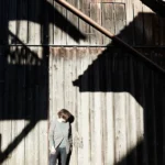 Gabi Veit steht im Sonnenlicht vor einer verwitterten Holzscheune, starke Schattenlinien fallen diagonal über die Fassade.