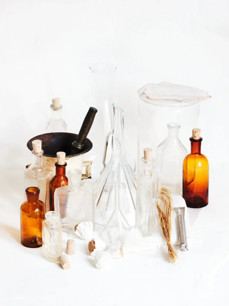 Verschiedene leere Glasflaschen in unterschiedlichen Formen, Größen und Farben (Klarglas und Bernstein), zusammen mit einem Mörser, Trichtern und Korken auf weißem Grund.