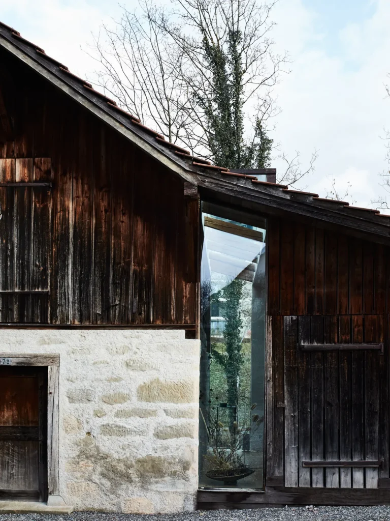 Außenansicht einer alten Holzscheune mit Steinwand und schmalem Glasfenster.