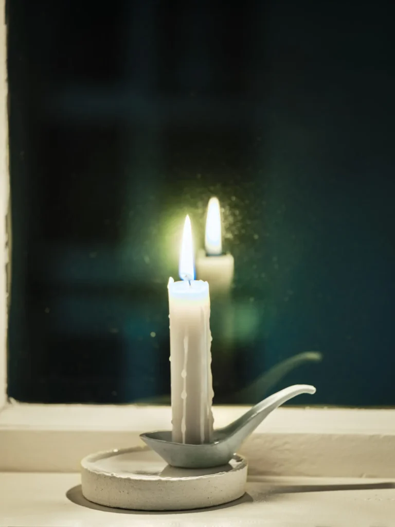 Weiße Kerze in kleinem Kerzenhalter auf Fensterbank, Flamme spiegelt sich im dunklen Glas.