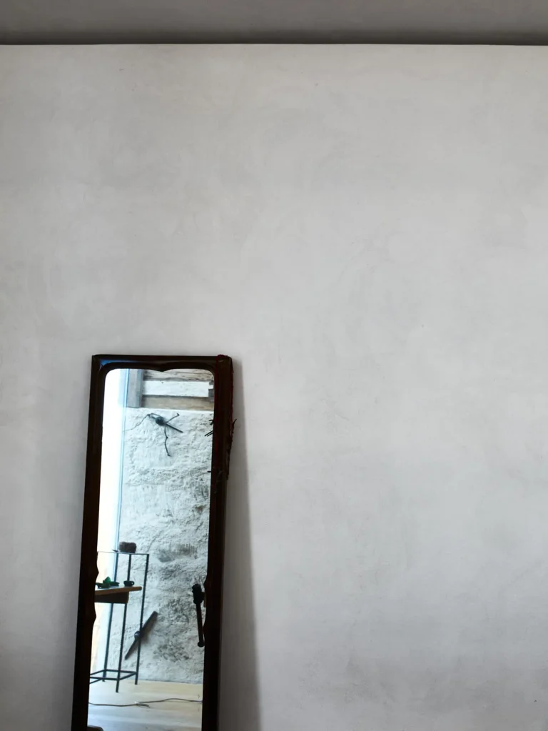 Schmaler Standspiegel vor weißer Wand, reflektiert Steinwand und kleines Regal.