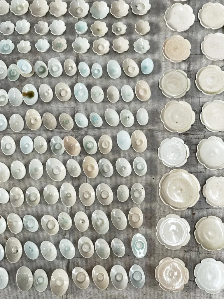 Reihenweise angeordnete kleine Keramikschalen und -schälchen in Weiß- und Pastelltönen, mit unterschiedlichen Glasuren und feinen Craquelé-Strukturen, auf grauem Untergrund ausgelegt.