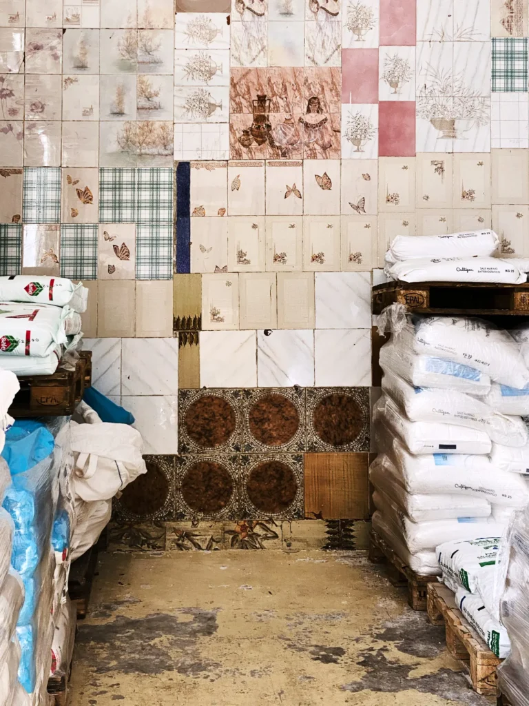 Patchwork-Wand aus unterschiedlich gemusterten Fliesen; davor Paletten mit weißen Säcken in einem Lagerraum