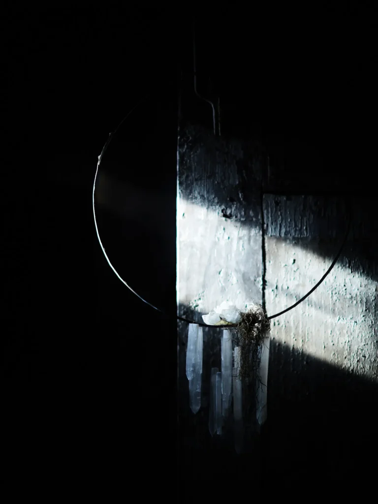 Metallring mit hängenden, klaren Kristallen vor einer rauen Wand; starkes Seitenlicht trifft die Kristalle, der Rest des Raums liegt im Dunkeln.