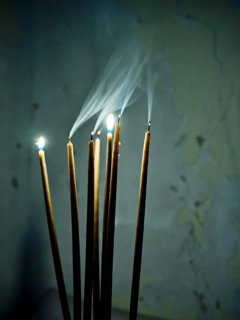 Schlanke Kerzen mit kleinen Flammen und aufsteigendem Rauch vor dunklem Hintergrund.