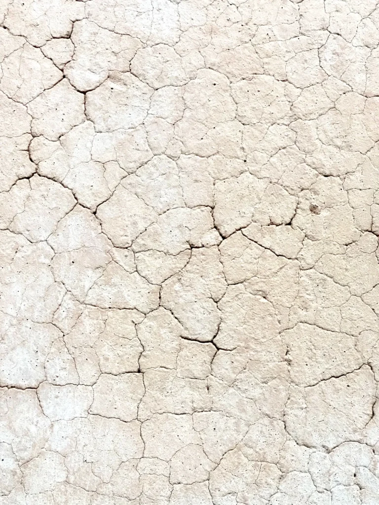 Nahaufnahme einer hellen, rissigen Oberfläche aus getrocknetem Lehm oder Kalk mit feinem, netzartigem Muster