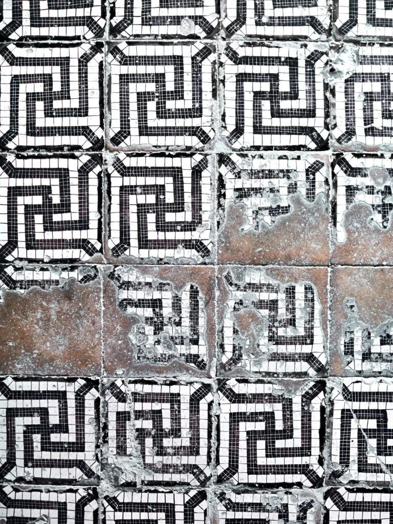 Abgenutzte schwarz-weiße Mosaikfliesen mit geometrischem Labyrinthmuster, stellenweise beschädigt und verwittert
