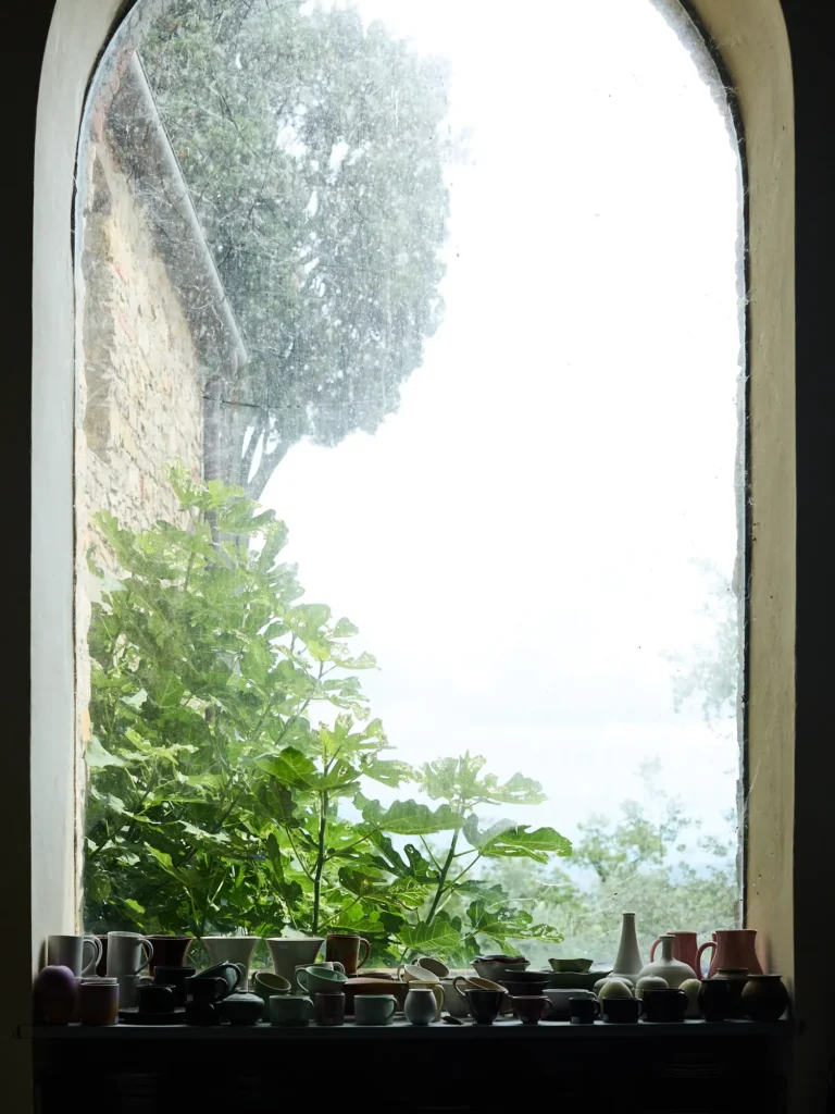 Keramikbecher und kleine Gefäße auf einer Fensterbank vor einem großen, gewölbten Atelierfenster mit Blick ins Grüne