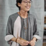 Porträtaufnahme einer lächelnden Frau, Rumi, mit Brille in grauem Leinenkleid, die Essstäbchen in den Händen hält, fotografiert von Dietlind Wolf vor hellem, textilem Hintergrund.