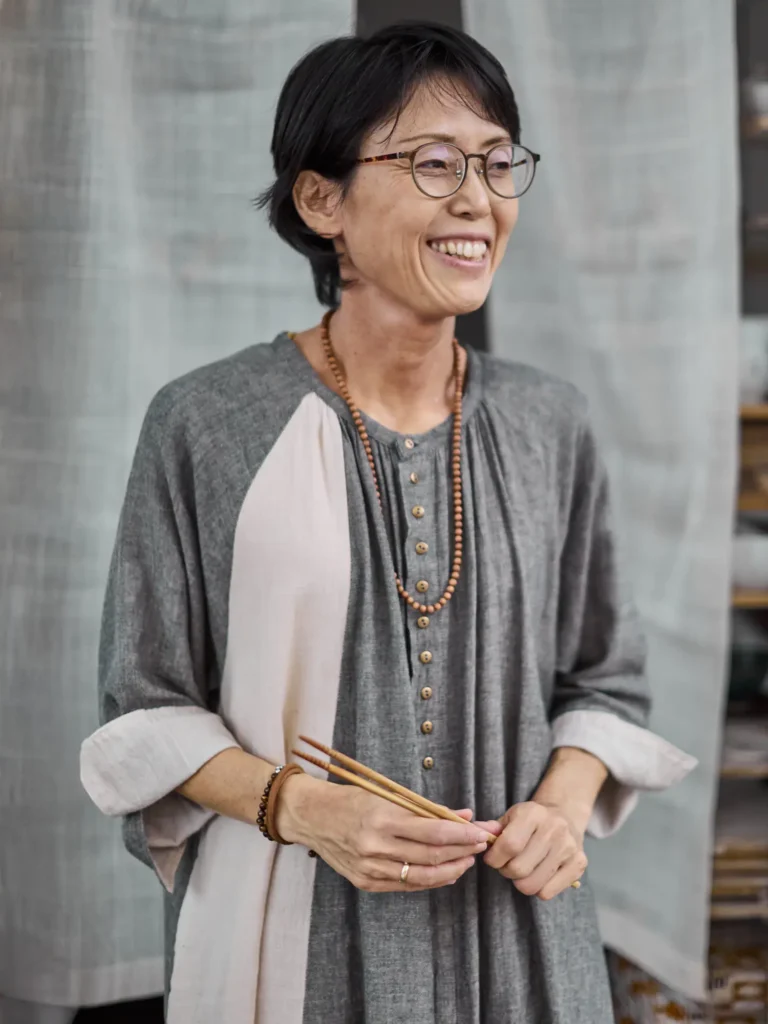 Porträtaufnahme einer lächelnden Frau, Rumi, mit Brille in grauem Leinenkleid, die Essstäbchen in den Händen hält, fotografiert von Dietlind Wolf vor hellem, textilem Hintergrund.