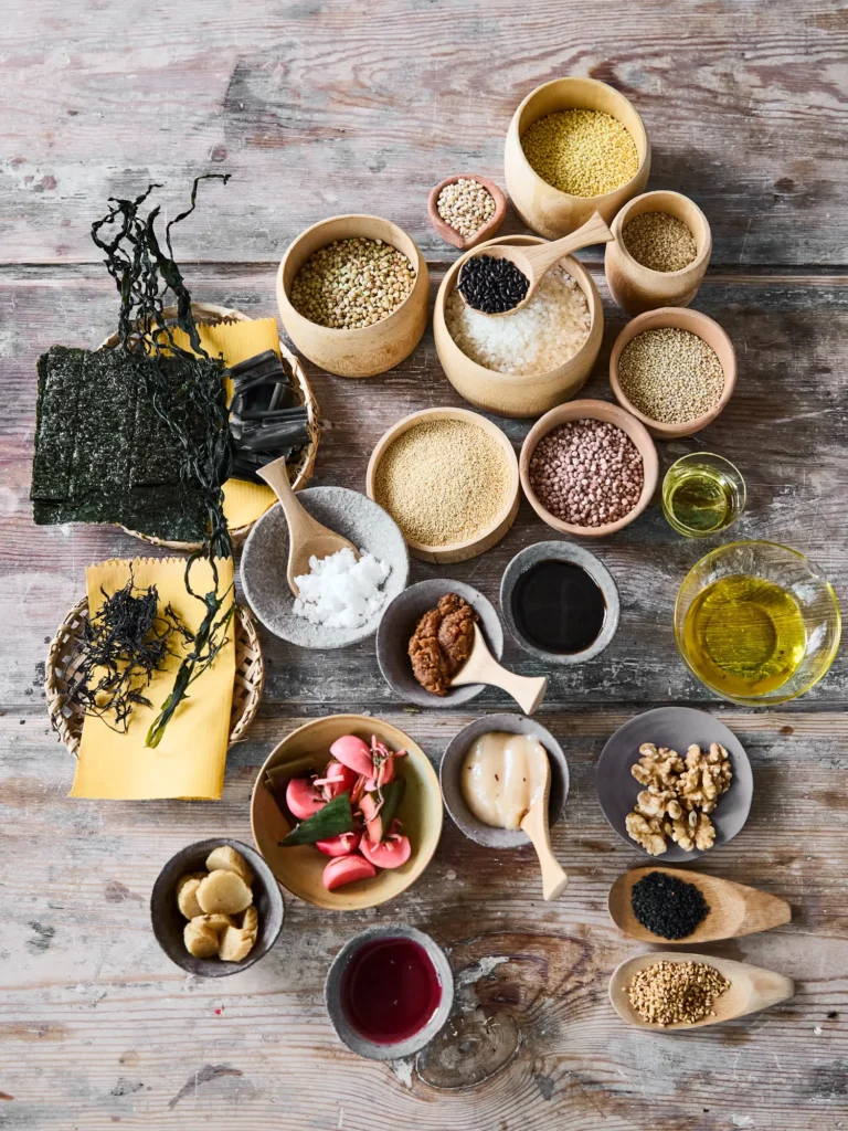 Vielfältige Anordnung kleiner Schalen mit Getreide, Hülsenfrüchten, Algen, Nüssen und Pasten sowie Öl in Glasschalen von Dietlind Wolf, arrangiert auf einer rustikalen Holzoberfläche.