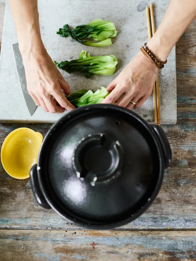 Draufsicht auf Hände beim Arrangieren von gedünstetem Pak Choi auf einem grauen Steinbrett neben einem schwarzen Tontopf und Essstäbchen.