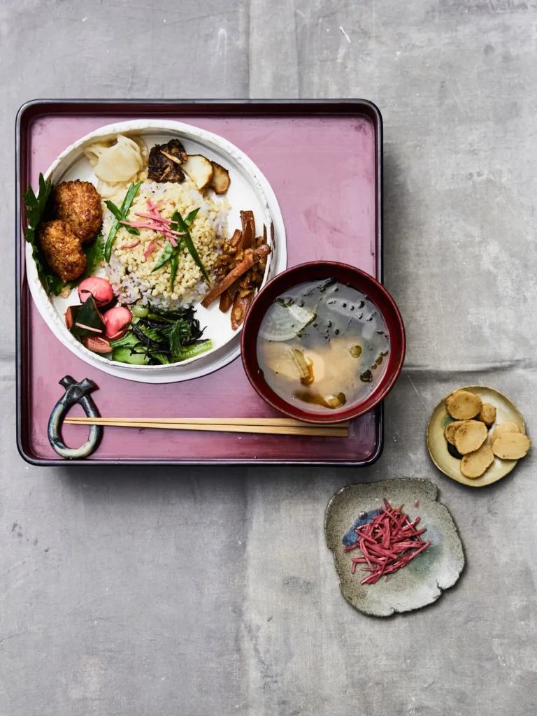 Japanisches Lunch-Ensemble auf einem weinroten Tablett, bestehend aus einer Schüssel Miso-Suppe und einem Teller mit Reis, Bratlingen und verschiedenen Pickles neben kleinen Beistelltellern, auf grauem Grund.