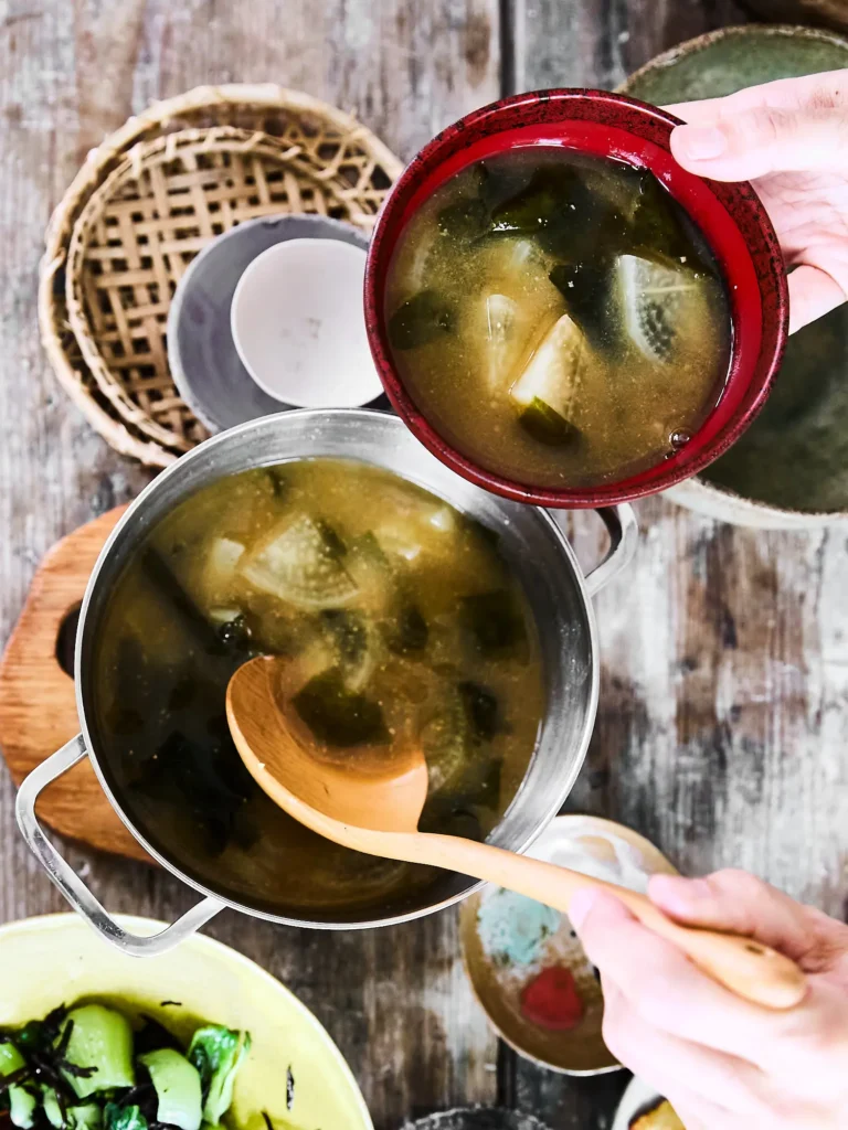 Hände halten eine Schale Miso-Suppe mit Daikon-Scheiben und Algen über einen großen Edelstahltopf mit Schöpfkelle von Dietlind Wolf, auf rustikalem Holzgrund.