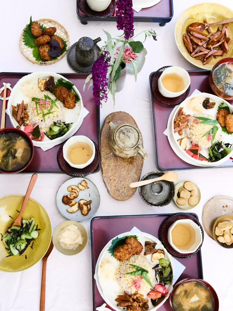 Gedeckter Tisch mit mehreren japanischen Lunch-Tabletts, einer Teekanne auf einem Steinbrett, Schüsseln mit grünem Gemüse und einer Blumenvase mit lila Blüten von Dietlind Wolf, in der Draufsicht.