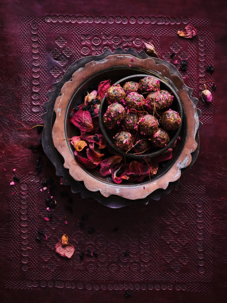 Dunkle Bliss Balls in einer flachen, rötlichen Keramikschale, bestreut mit getrockneten Rosenblüten und hellen Samen, daneben eine kleine Schale mit getrockneten Rosenknospen und ein Häufchen heller Samen von Dietlind Wolf, auf grauem Grund.