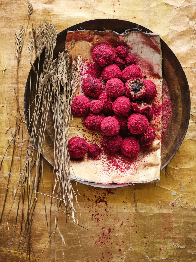 Rötliche Blissballs mit dunkler Füllung auf einem braunen Keramikteller, umgeben von Weizenähren, auf hellbraunem Hintergrund.