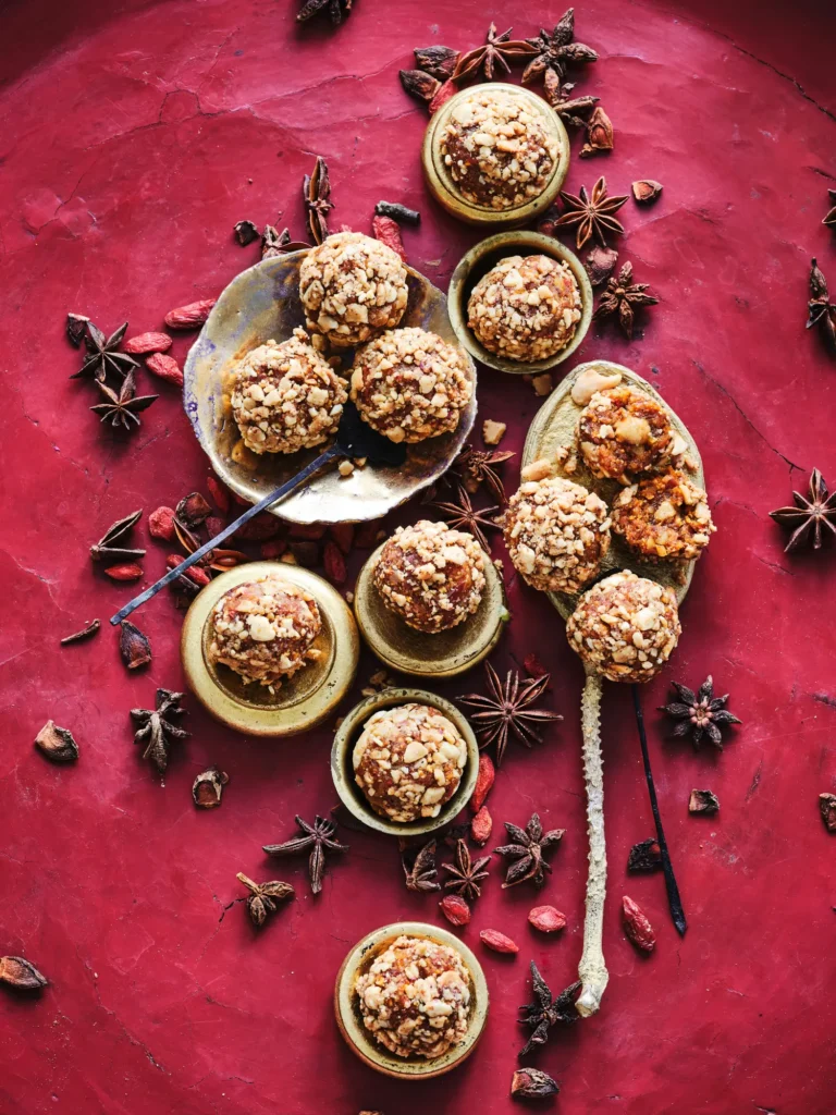 Braune Bliss Balls, angeordnet in kleinen Schälchen und bestreut mit Sesam sowie hellen Samen, daneben Gojibeeren und Gewürze von Dietlind Wolf, auf rotem Hintergrund.