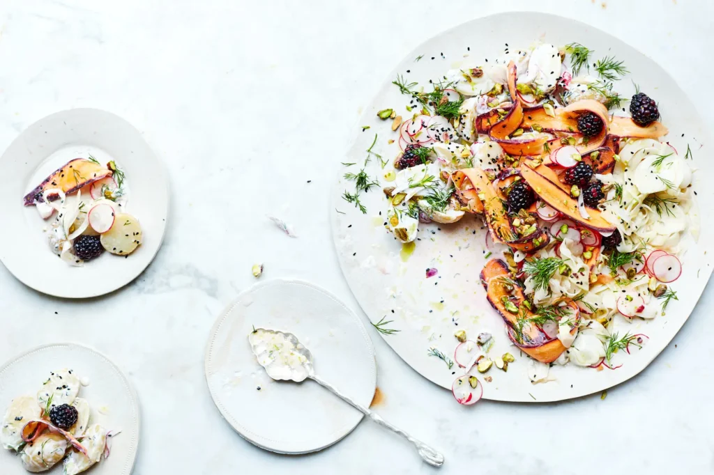 Moderner Kartoffelsalat auf hellen Tellern, garniert mit violetten Karottenstreifen, Radieschen, Brombeeren, Dill und gehackten Pistazien auf einer Marmoroberfläche.