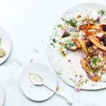 Moderner Kartoffelsalat auf hellen Tellern, garniert mit violetten Karottenstreifen, Radieschen, Brombeeren, Dill und gehackten Pistazien auf einer Marmoroberfläche.