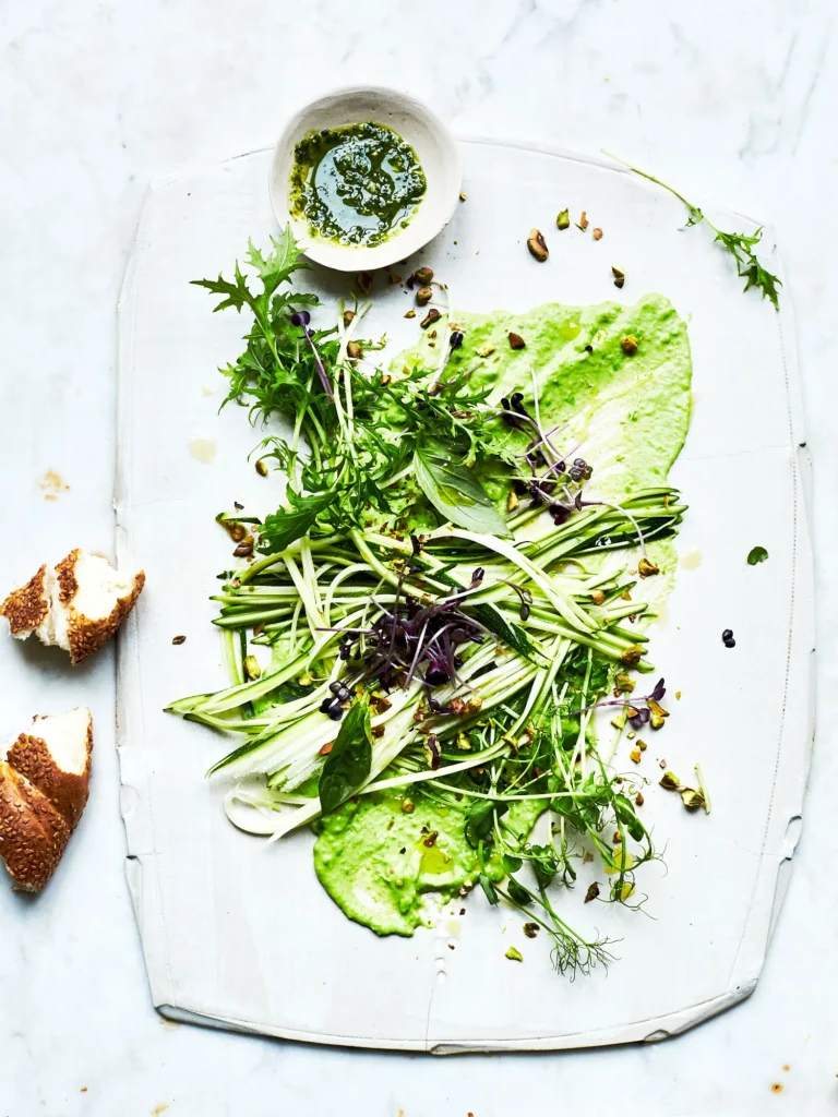 Moderne Gemüseplatte mit hellgrünem Püree, Zucchini-Zoodles, frischen Microgreens und gehackten Pistazien, serviert mit Kräuter-Pesto und Sesambrot.
