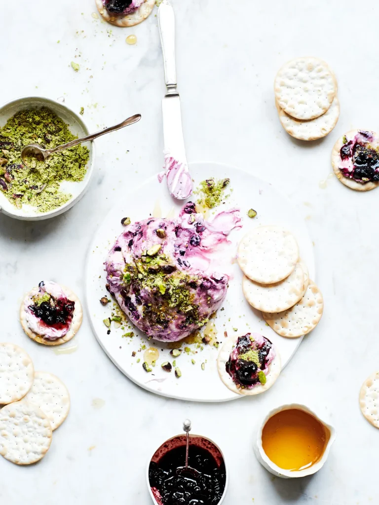 Labneh-Dip mit Beeren, Honig und Pistazien auf Crackern angerichtet, umgeben von kleinen Schälchen mit Zutaten auf Marmor.