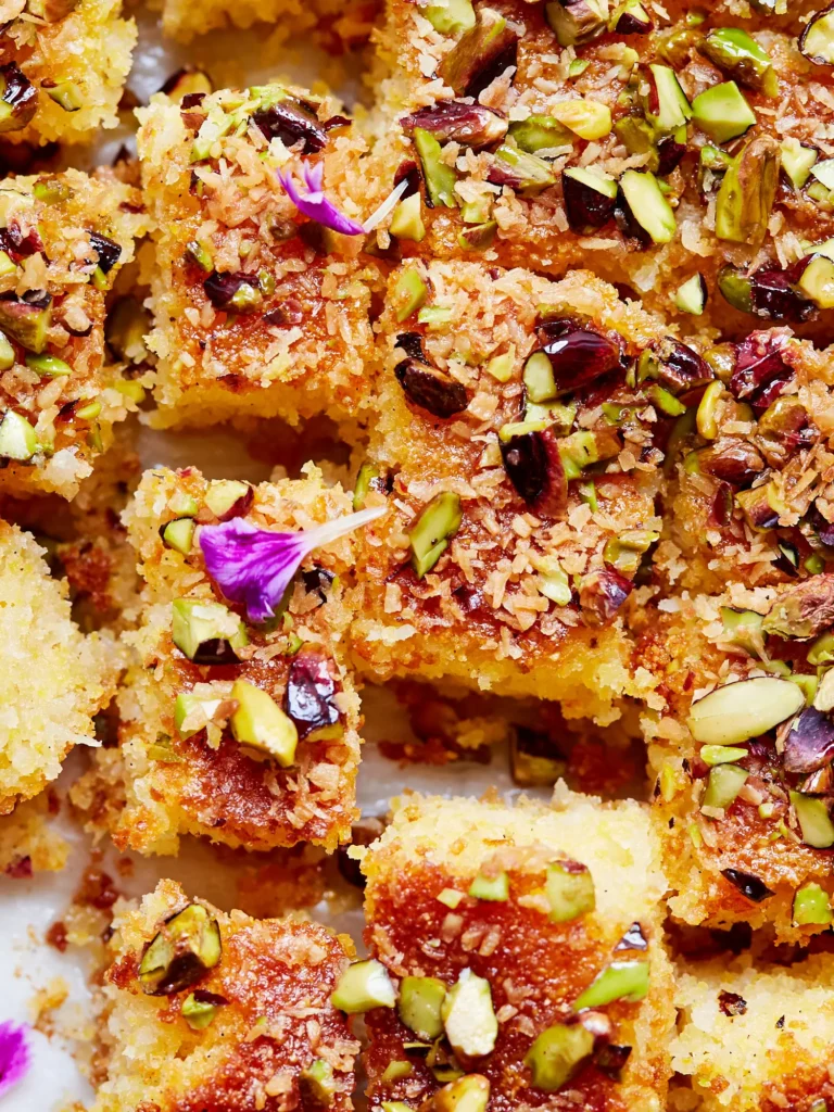 Saftige, goldbraune Grießkuchenstücke mit Sirup, gehackten Pistazien und Kokosraspeln, dekoriert mit kleinen violetten Blüten.