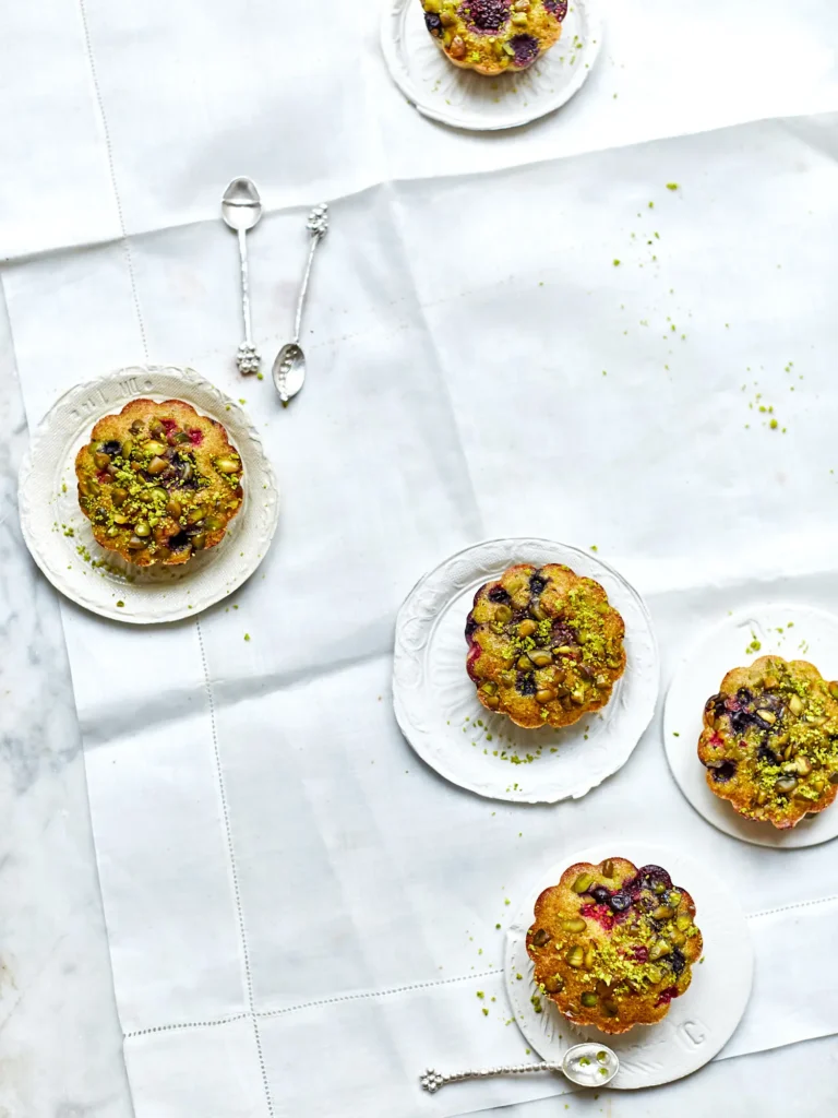 Kleine, goldbraune Küchlein in Blumenform, gebacken mit Beeren und großzügig mit Pistazienstaub bestreut.