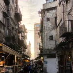 Enge Altstadtgasse mit Marktständen, alten Wohnhäusern und Passanten im warmen Abendlicht.