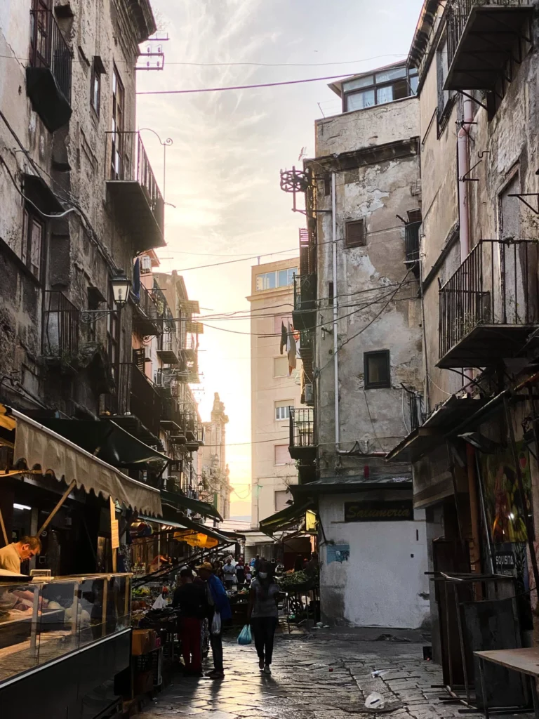 Enge Altstadtgasse mit Marktständen, alten Wohnhäusern und Passanten im warmen Abendlicht.